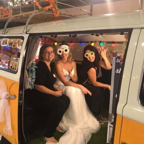 Trois femmes sont assises dans une camionnette vintage aux couleurs vives. La femme au milieu porte une robe blanche et des lunettes en forme de fleur. Les deux autres femmes sourient également et portent des lunettes de fête. L'intérieur de la camionnette est orné de lumières et de décorations colorées.