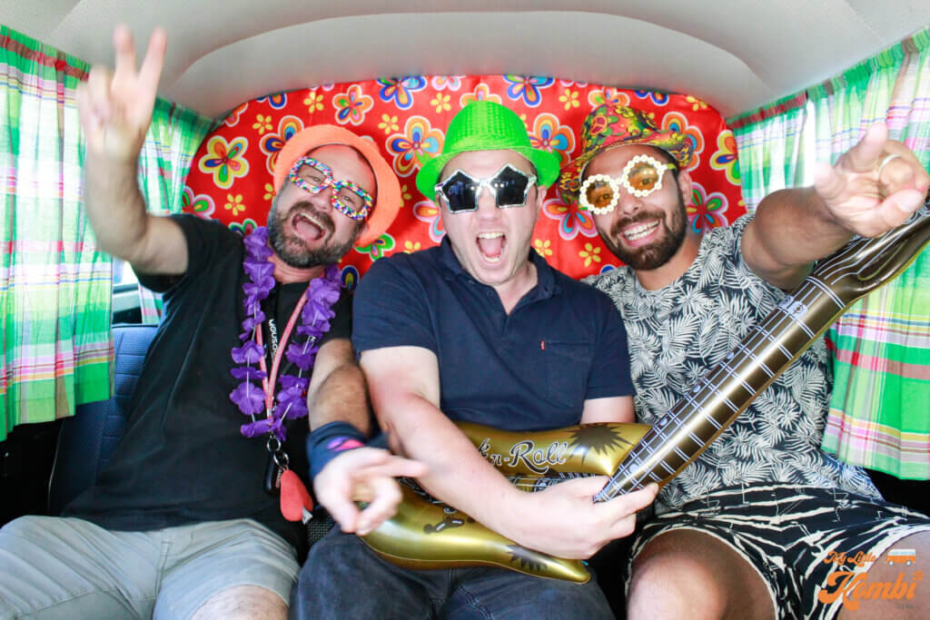 Trois hommes s'amusent dans une camionnette colorée et décorée. Ils portent des accessoires de fête : un chapeau vert, des lunettes en forme d'étoile, un boa à plumes et des lunettes de soleil fantaisie. Un homme tient une guitare gonflable et ils semblent tous applaudir et sourire.