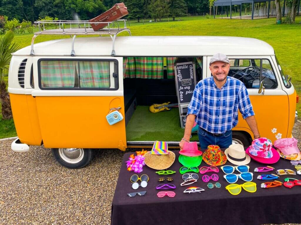 Un homme vêtu d'une chemise à carreaux bleu et blanc et d'un chapeau gris se tient à côté d'une camionnette vintage jaune avec une table devant lui affichant une gamme de chapeaux et de lunettes de soleil colorés. La camionnette a une valise sur le toit et des portes ouvertes révélant un intérieur vert.