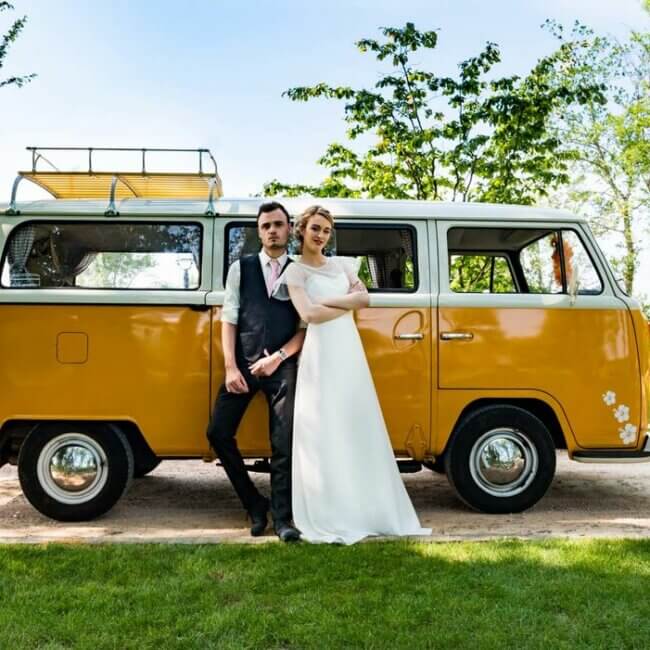 Un couple s’appuie contre une camionnette Volkswagen jaune vintage dans un cadre extérieur pittoresque. L'homme, vêtu d'un costume, se tient debout, une main dans la poche. La femme, vêtue d’une robe blanche fluide, se tient debout, les bras croisés. Des arbres et de l'herbe verte les entourent.