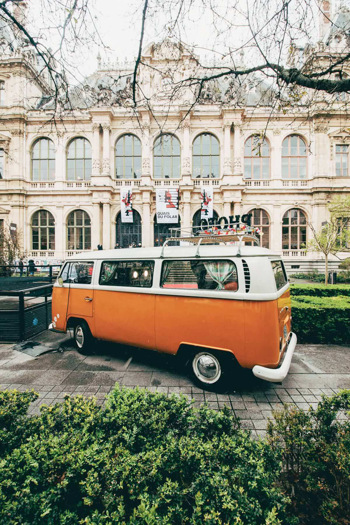 Une camionnette orange vintage est garée dans une rue pavée devant un bâtiment orné de fenêtres cintrées et de détails architecturaux complexes. La camionnette dispose d'une galerie de toit avec bagages. Autour de la scène se trouvent des arbres et de la verdure.