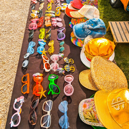 Une table noire du Domaine des marais Bourgoin présente de nombreuses lunettes de soleil et chapeaux colorés. Les lunettes de soleil sont disponibles dans différentes formes et couleurs, notamment des montures en forme de cœur et des nuances de jaune, rose, vert et bleu. Les chapeaux comportent des casquettes dorées brillantes, des embellissements de paillettes et des motifs floraux.