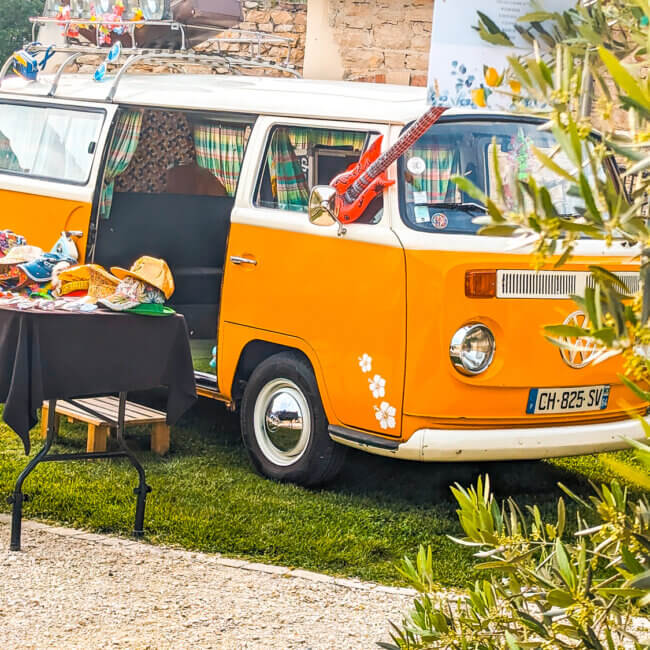 Une camionnette orange vintage colorée avec sa porte latérale ouverte est garée à côté d'une table présentant divers chapeaux et accessoires au Domaine des marais Bourgoin. La camionnette, décorée d'autocollants et dotée d'une galerie de toit, est encadrée d'arbres et d'arbustes.