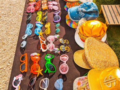 Une table noire du Domaine des marais Bourgoin présente de nombreuses lunettes de soleil et chapeaux colorés. Les lunettes de soleil sont disponibles dans différentes formes et couleurs, notamment des montures en forme de cœur et des nuances de jaune, rose, vert et bleu. Les chapeaux comportent des casquettes dorées brillantes, des embellissements de paillettes et des motifs floraux.